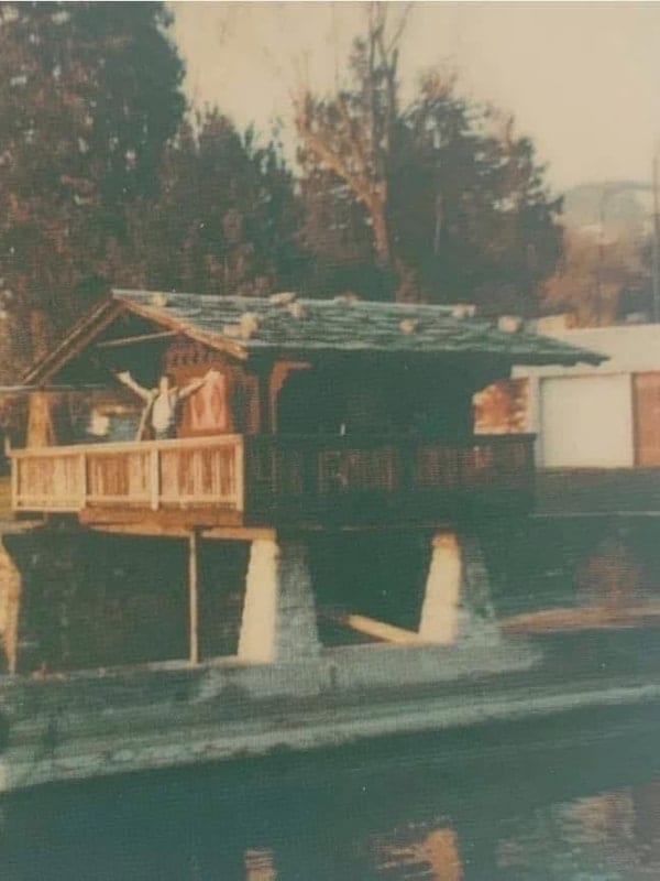 Freddie Mercury mit erhobenen Armen auf dem kleinen Bootshaus am See der Duck House, der von ihm gemieteten Villa in Clarens.